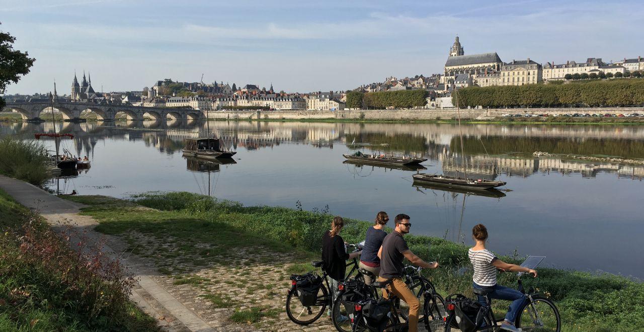 Group of cyclists by the river, with a view of a historic city and boats.