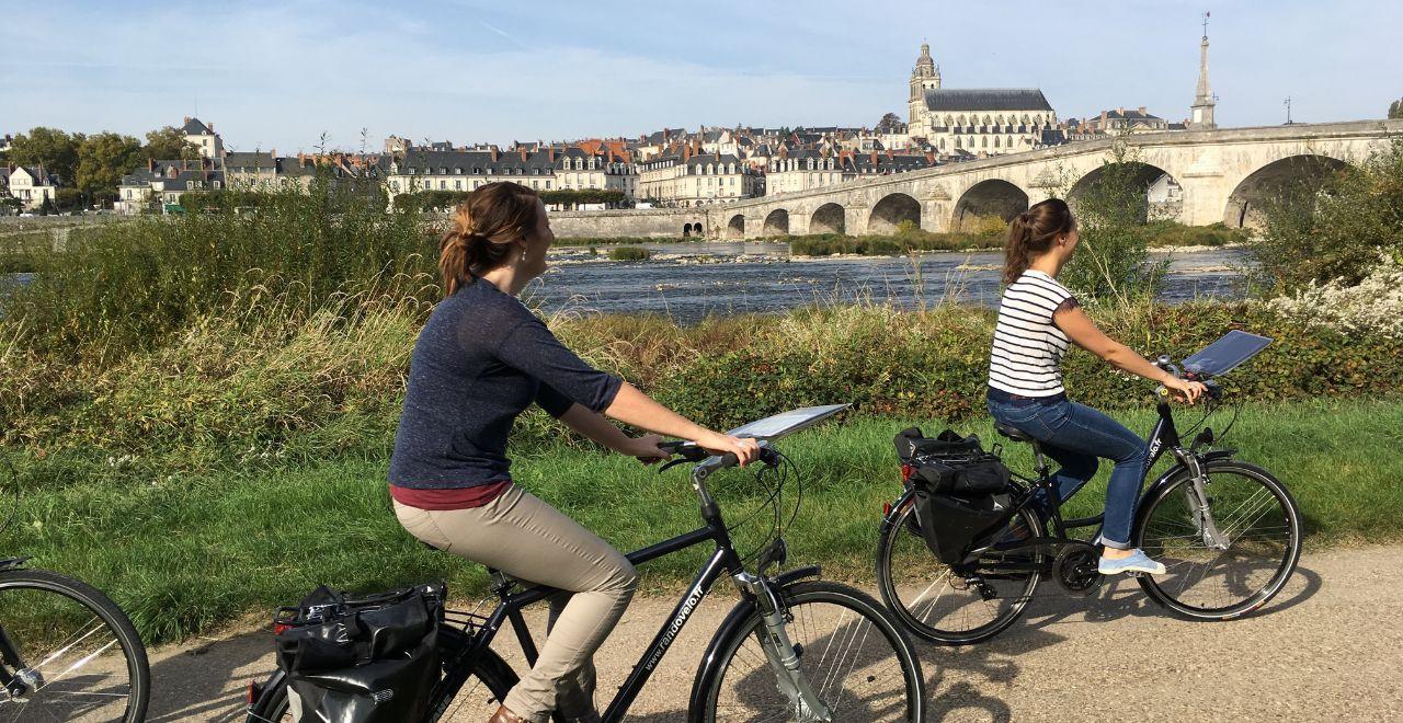 Two women cycling along a river with a historic city and bridge in the background.