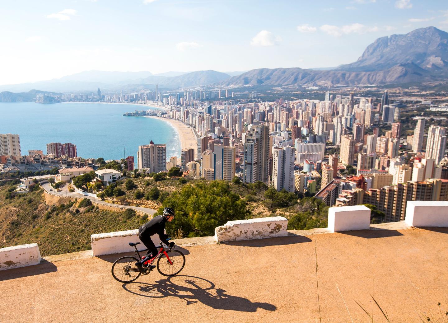 “Aerial view of cyclists riding on a winding road with a vast cityscape, coastline, and mountains in the background in Calpe, Costa Blanca.”