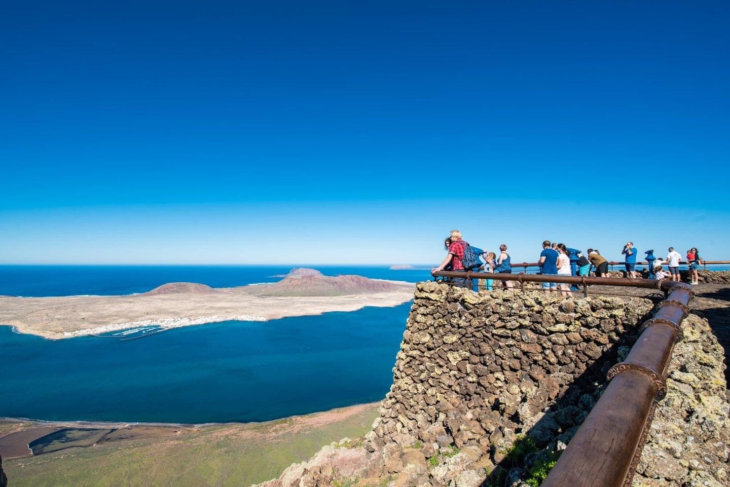 Tourists enjoying the scenic view from the Mirador del Río viewpoint in Lanzarote, overlooking the ocean and La Graciosa island under a clear blue sky.