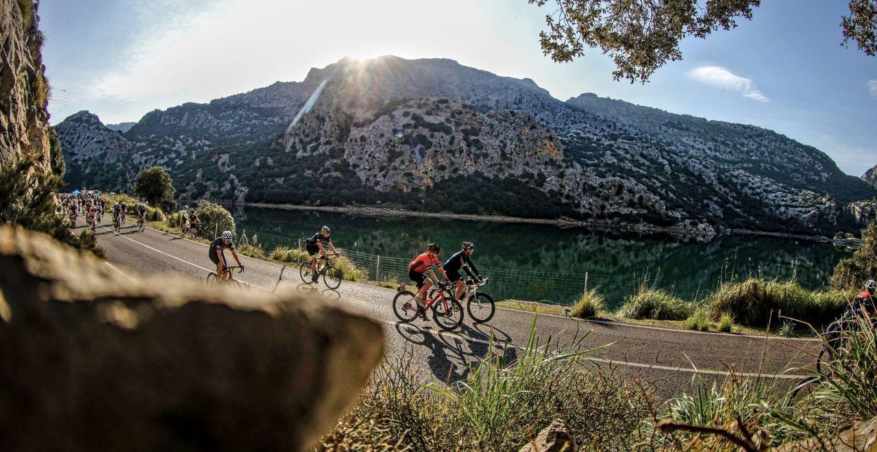 Cyclists riding along a lakeside mountain road at sunset.
