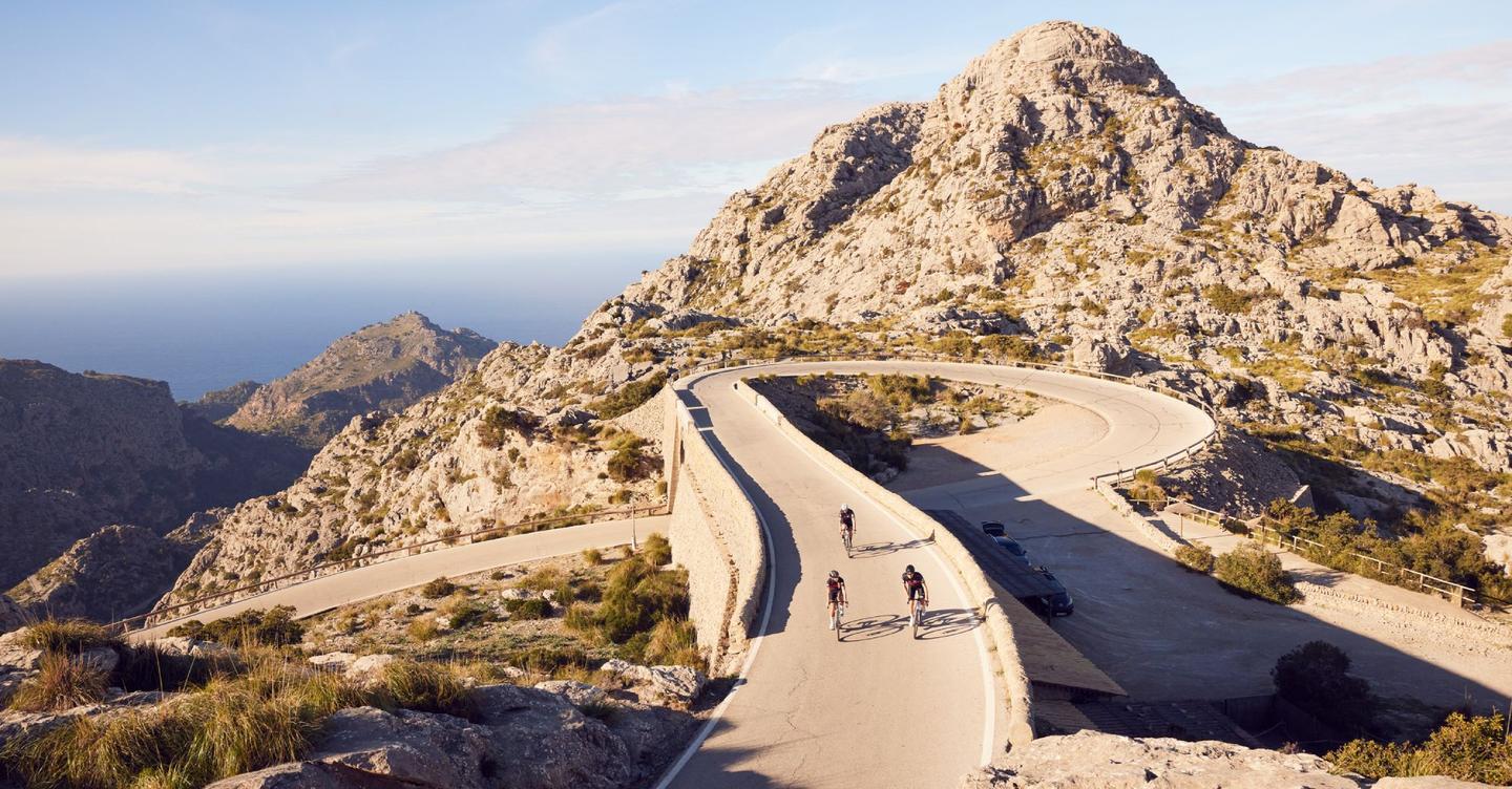 Three cyclists navigating the winding roads of Coll de Soller in Mallorca, surrounded by steep, rocky mountains and lush greenery under a clear sky.