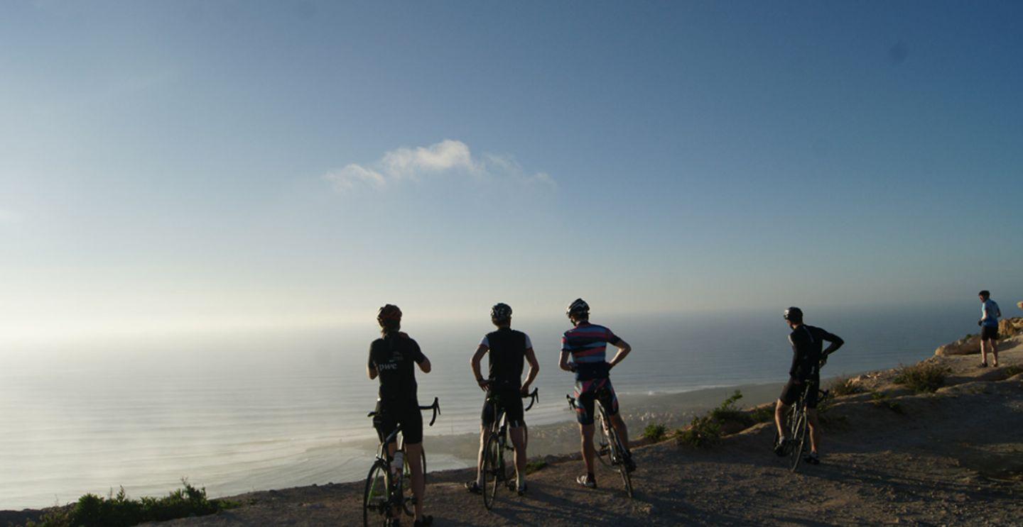 Cyclists standing on a hilltop, looking out over a misty view of the sea and coastline below.