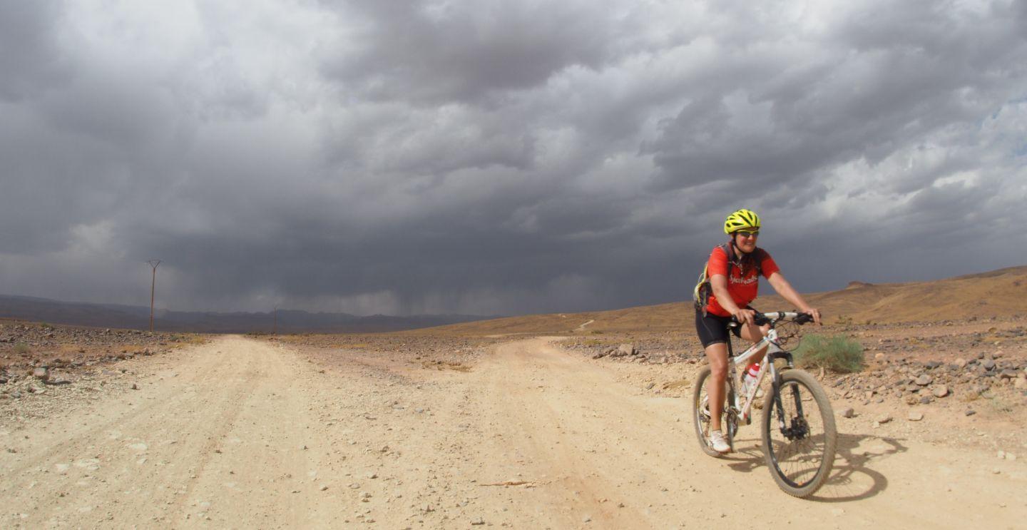 Cyclist riding on a dirt road under a cloudy sky, with mountains visible in the distance.