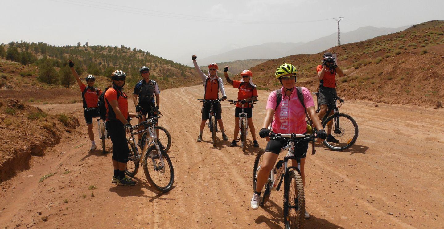 Group of cyclists on a dusty trail in a desert landscape, some raising their arms in celebration, with hills in the background.