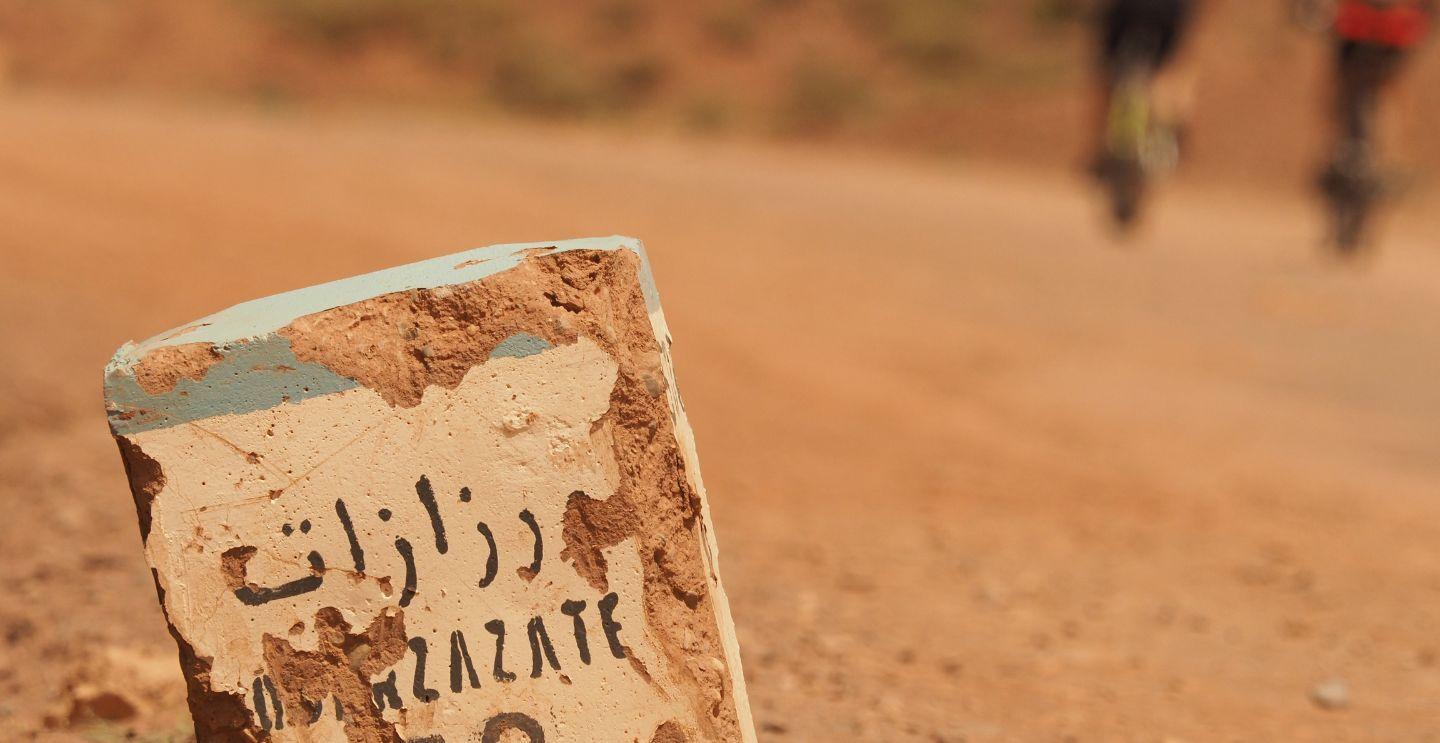 Weathered milestone with Arabic script marking the distance to Ouarzazate, on a dusty desert road.