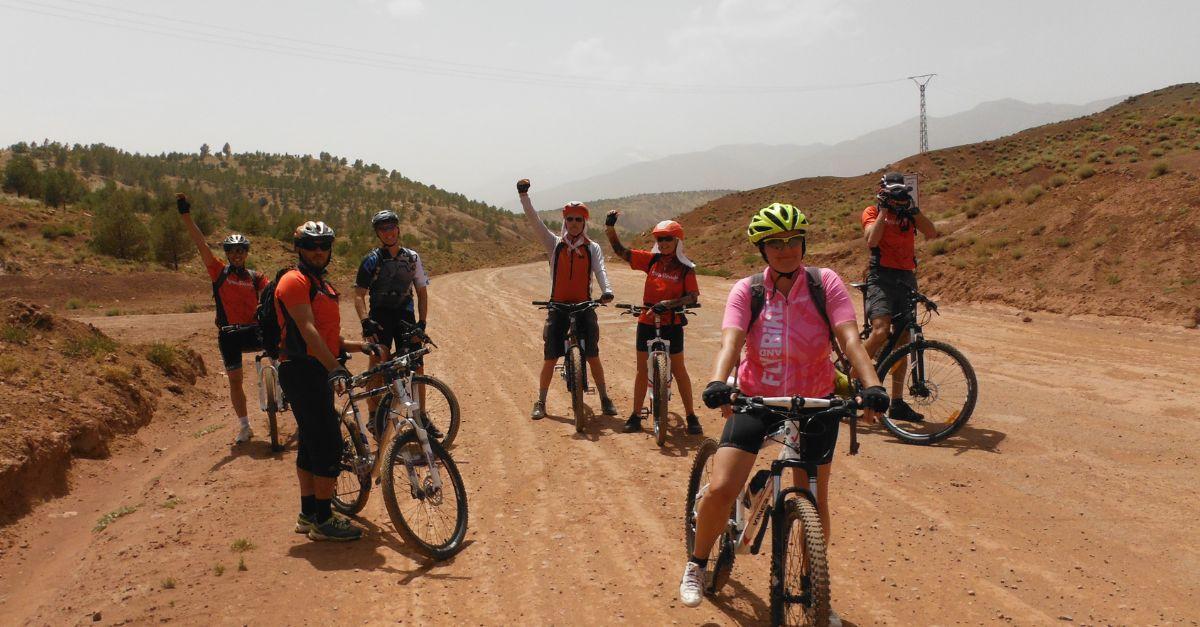 A group of mountain bikers on a dirt road with arid mountains in the background and a clear sky above in a Moroccan landscape