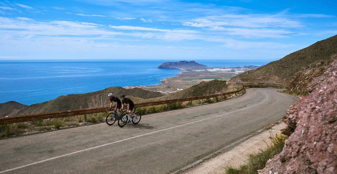 Two cyclists ride on a winding coastal road with a stunning view of the blue sea and distant mountains under a clear sky