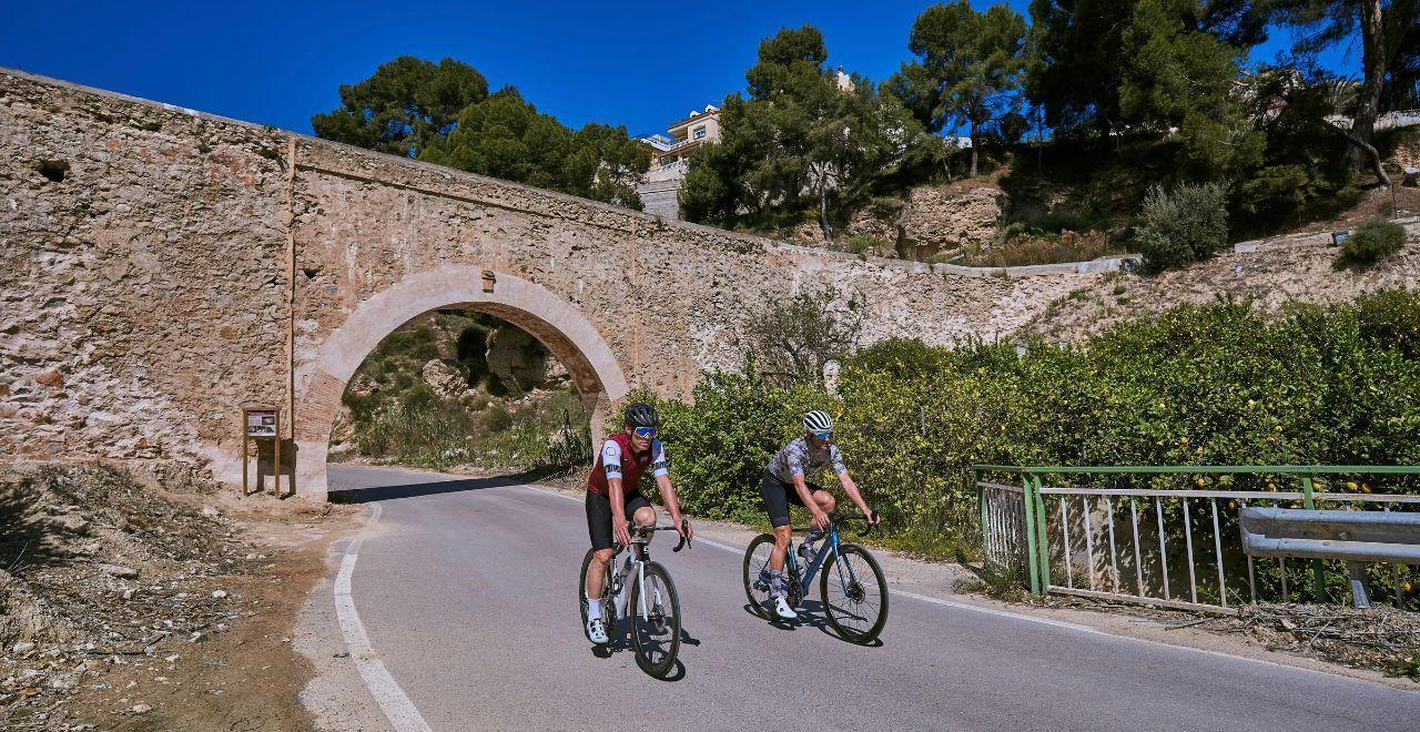  Two cyclists pass under an ancient stone arch bridge on a sunny day, with greenery and trees in the background.