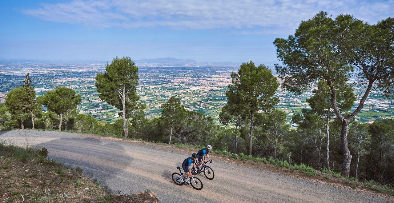 wo cyclists climb a mountain road, overlooking a vast cityscape and green fields with a clear blue sky above