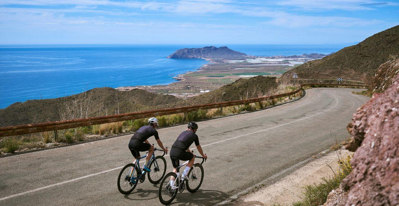  Two cyclists ascend a winding road with a beautiful coastal view, featuring a blue sea and rugged mountains under a clear sky.