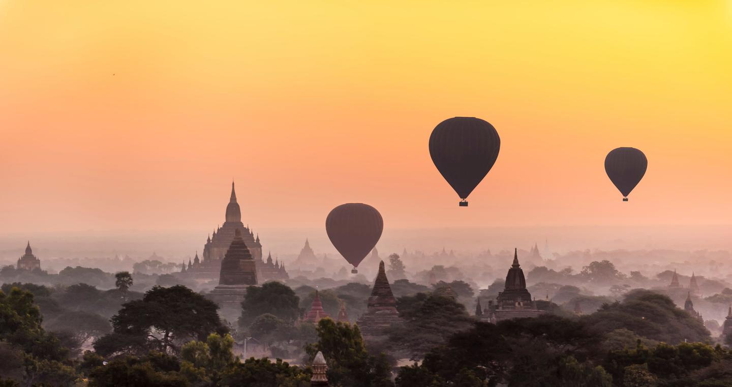 Image of hot air balloons floating over ancient temples and pagodas at sunrise in Bagan, Myanmar, with an orange and yellow sky.