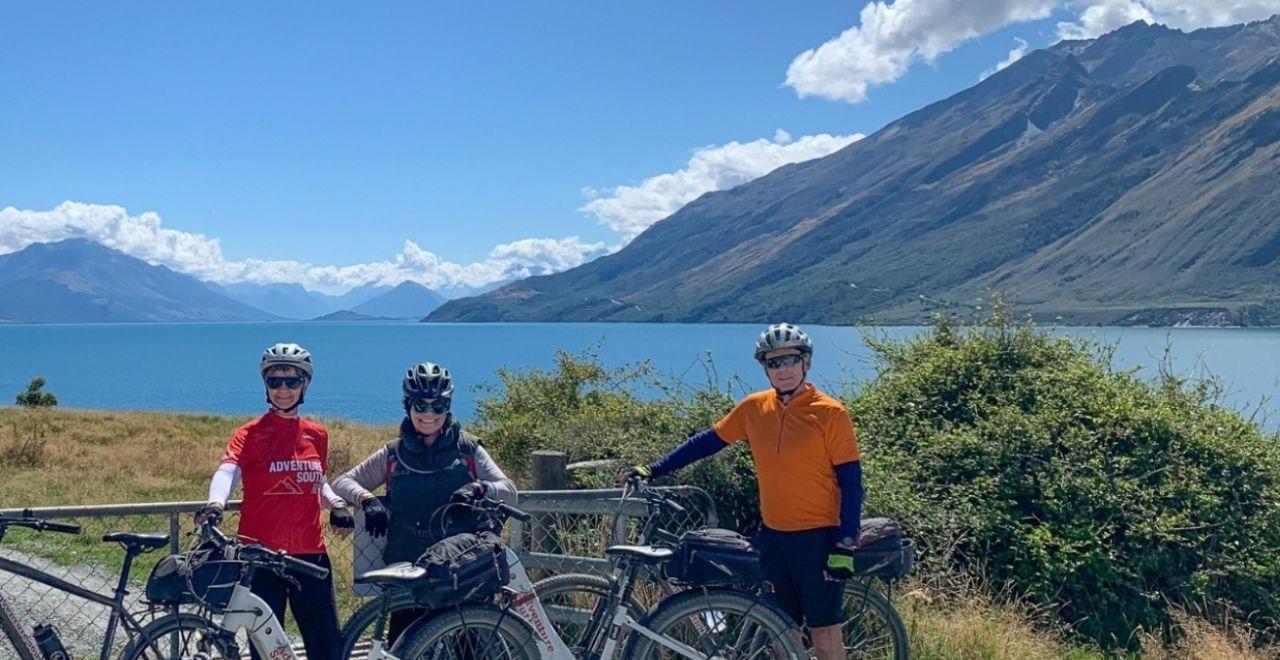 Three cyclists posing with bikes at Lake Hawea on the Alps 2 Ocean Cycle Trail.