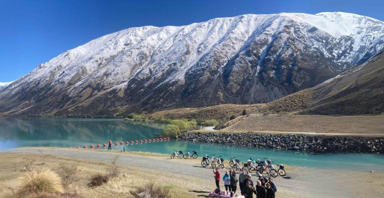 Group of cyclists by Lake Ohau with snow-capped mountains in the background on the Alps 2 Ocean Cycle Trail.