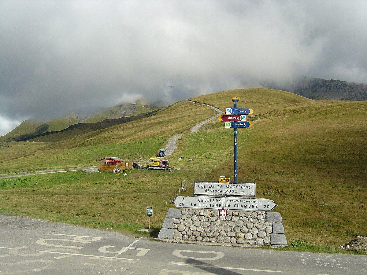 The summit sign of Col de la Madeleine at an altitude of 2000m, with various directional signs and a cloudy sky in the background.