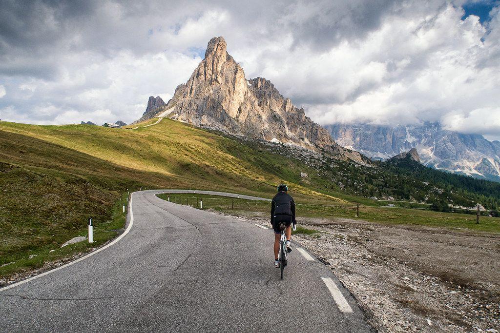 A cyclist rides along the winding road of Passo Giau in the Dolomites, with dramatic rocky peaks and green alpine meadows under a cloudy sky in the background.