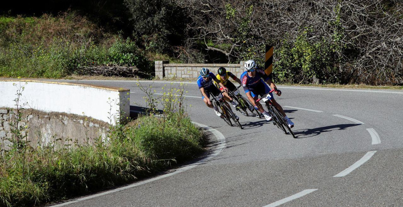 Three cyclists taking a sharp corner on a scenic countryside road, showcasing teamwork and skill.