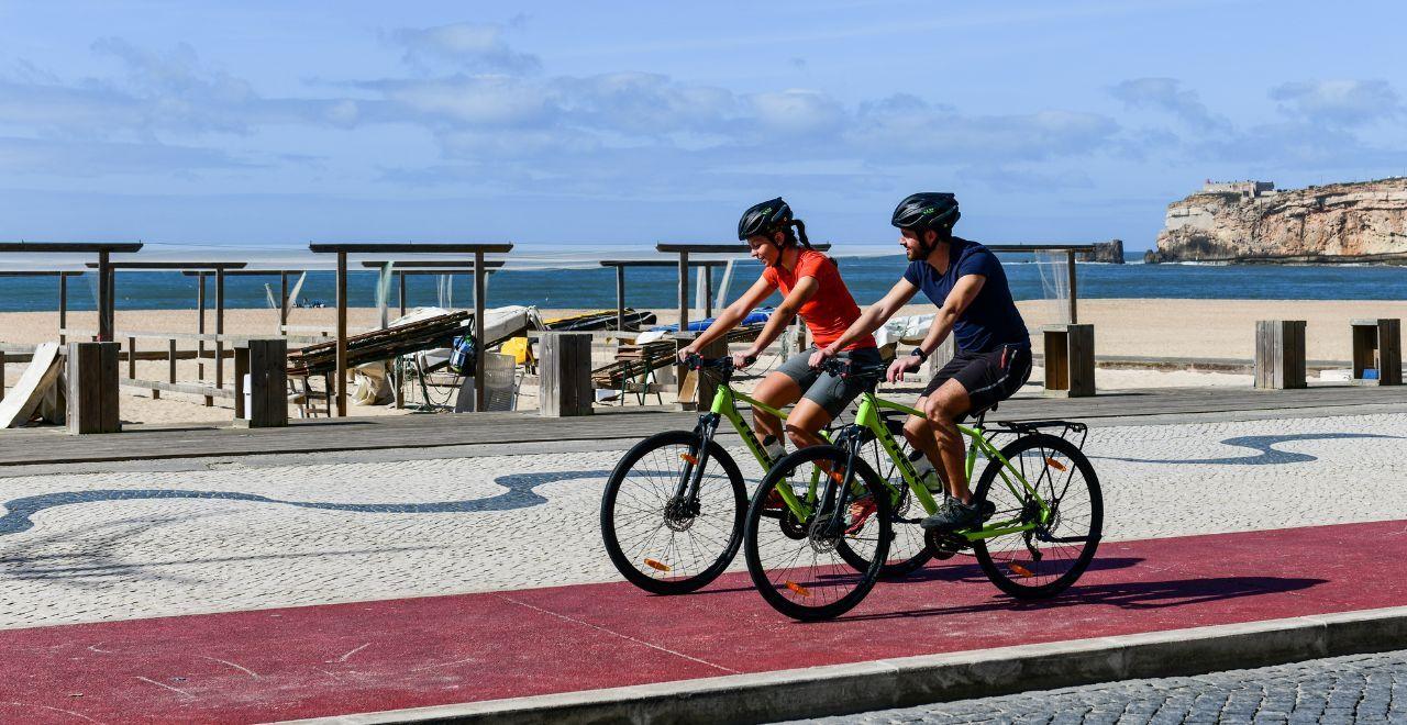 A man and a woman cycling side by side on a paved path by the beach with a coastal town in the background.