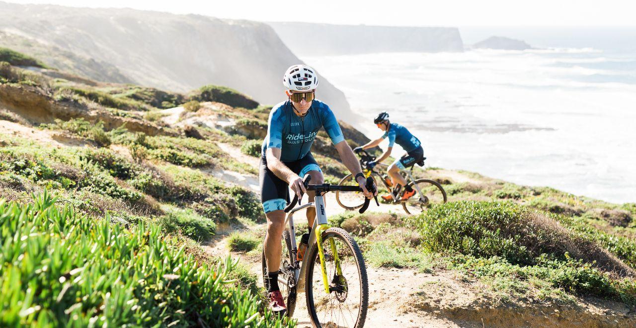 Two cyclists in blue jerseys navigating a rocky coastal trail with ocean waves in the background.