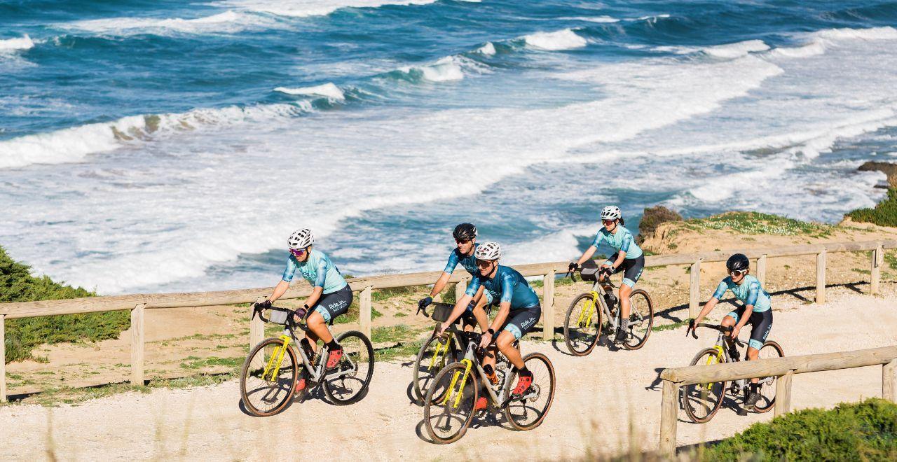 Group of cyclists in matching blue jerseys riding along a scenic coastal path with waves crashing in the background.