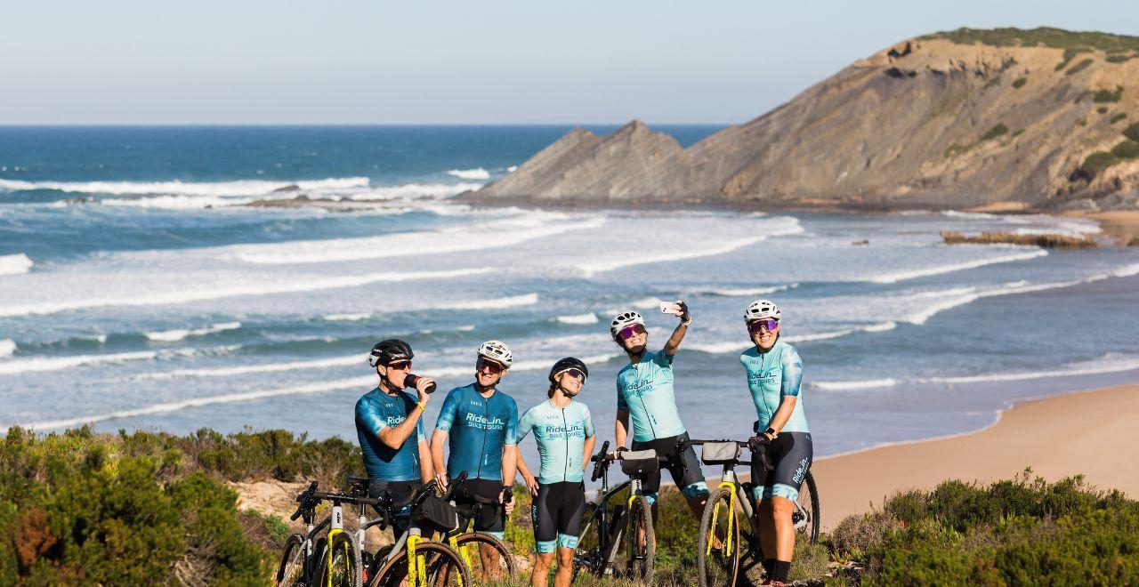 Group of cyclists in blue jerseys taking a break by the ocean, with one person taking a selfie.