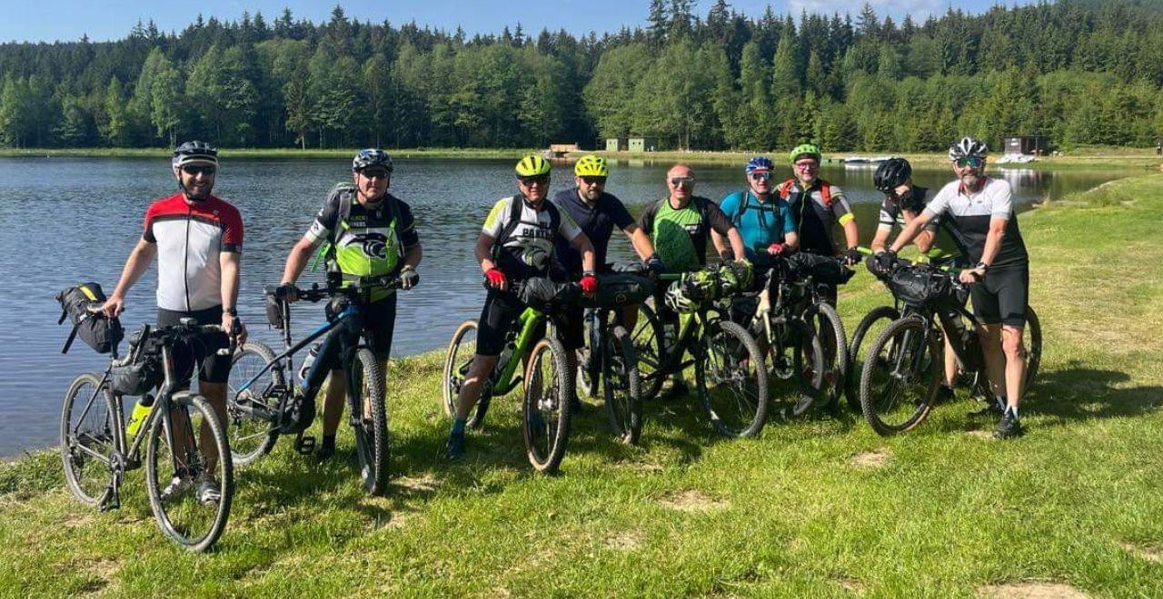 Group of cyclists posing with their bikes in front of a lake, surrounded by trees.