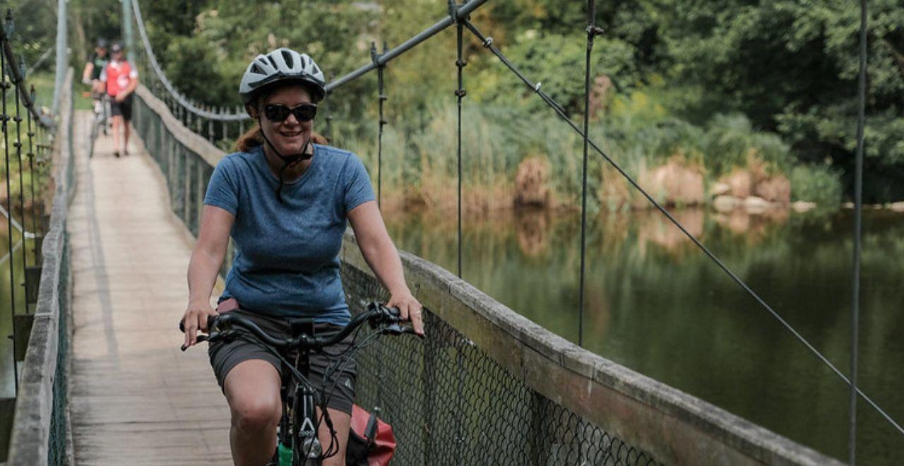 Female cyclist crossing a wooden suspension bridge over a river, smiling at the camera