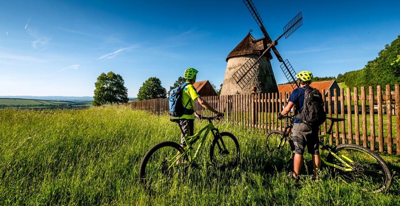 Two cyclists stopped in front of an old windmill surrounded by greenery.