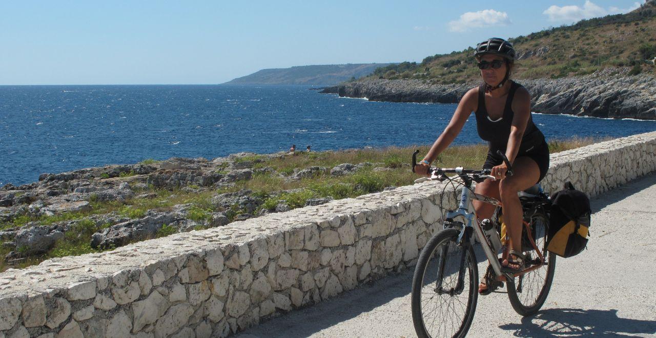 Woman cycling along a coastal path with rocky shoreline and blue sea.