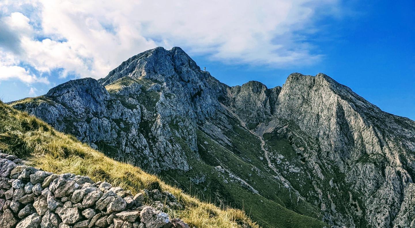Rugged mountain peaks of the Serra de Tramuntana in Mallorca, with a grassy slope and stone wall in the foreground, under a partly cloudy sky.