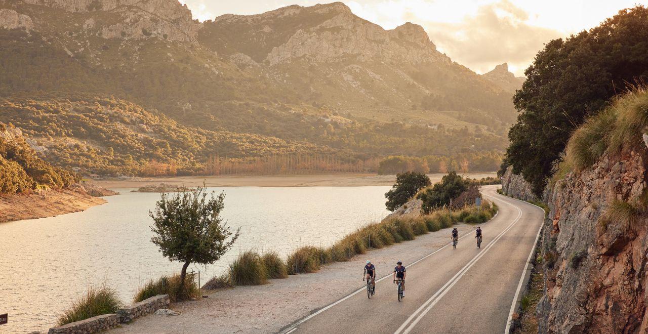 Four cyclists riding on a scenic mountain road near a lake at sunset