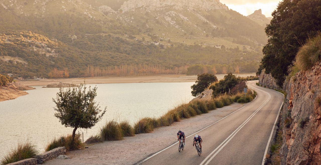 Two cyclists riding on a winding mountain road beside a lake at sunset