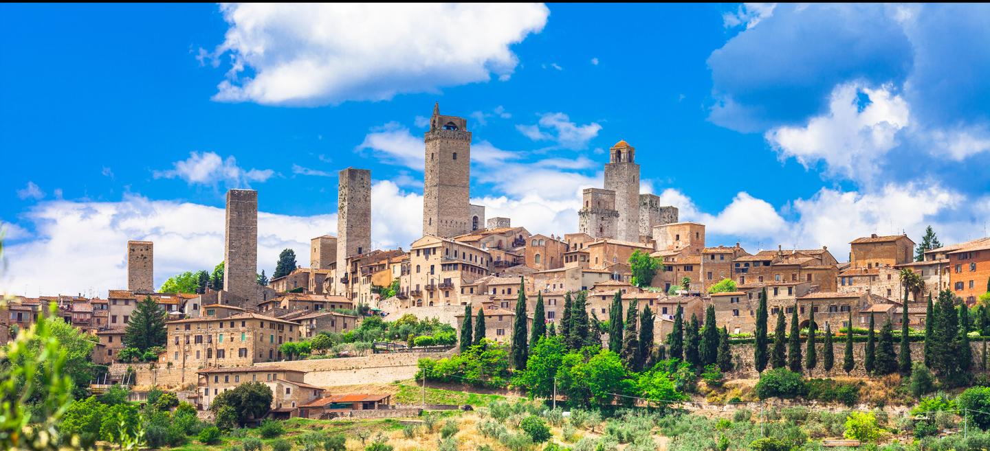 A picturesque view of San Gimignano, a historic town in Tuscany, Italy, featuring its iconic medieval towers, rustic buildings, lush greenery, and surrounding vineyards under a bright blue sky with scattered clouds.