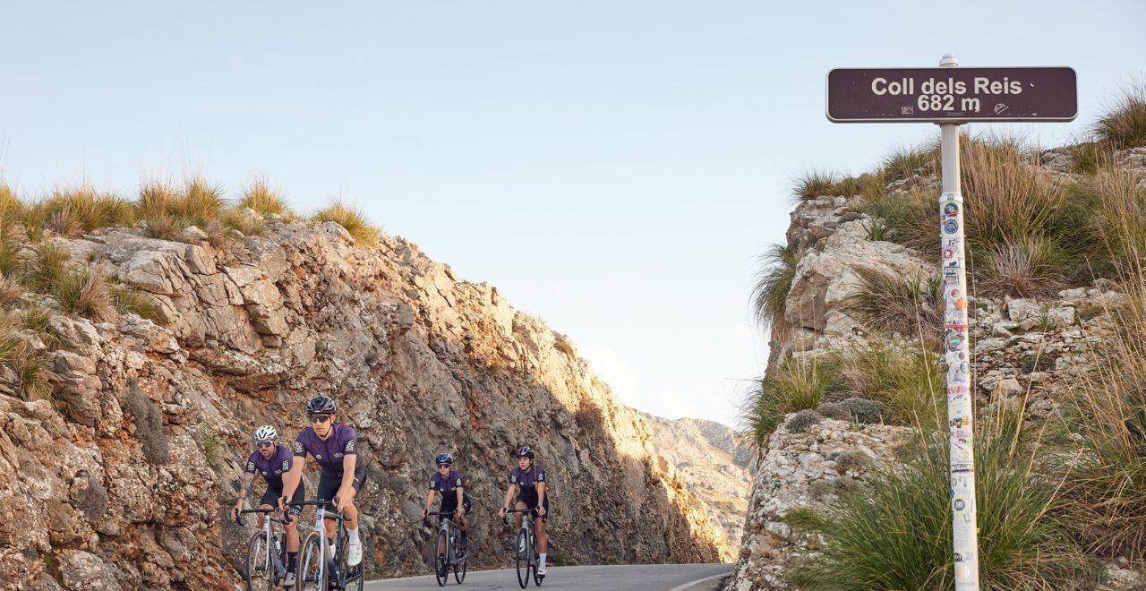 Cyclists passing the Coll dels Reis signpost on a winding road.