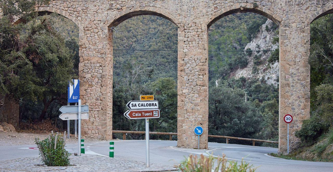 Road signs for Sa Calobra and Cala Tuent under a stone aqueduct in Mallorca.