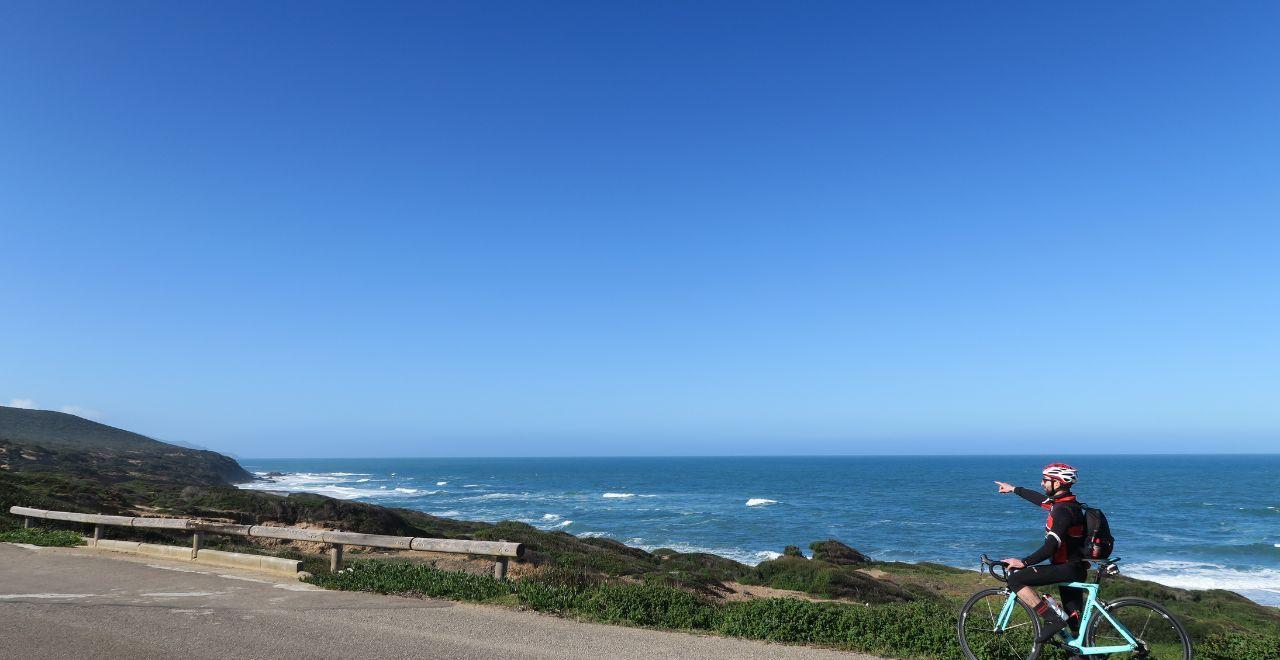 Cyclist pointing towards the ocean on a coastal road with clear blue skies.