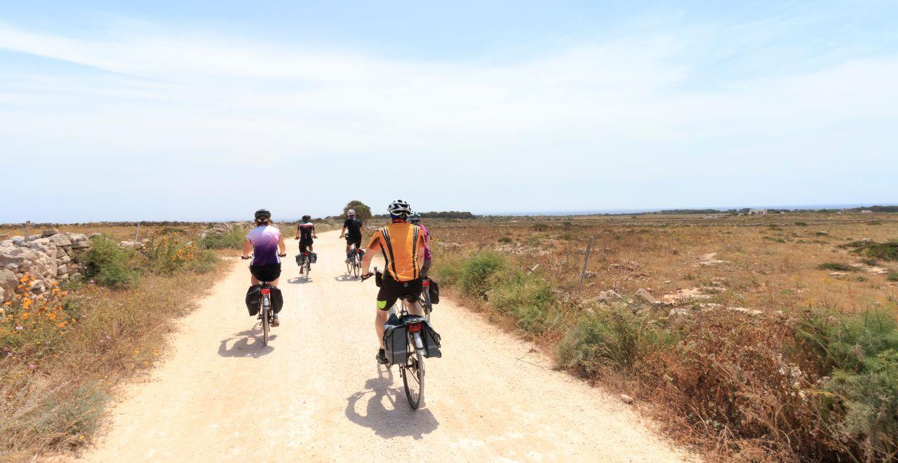 Cyclists on a dirt path through a dry, open landscape under a clear sky.