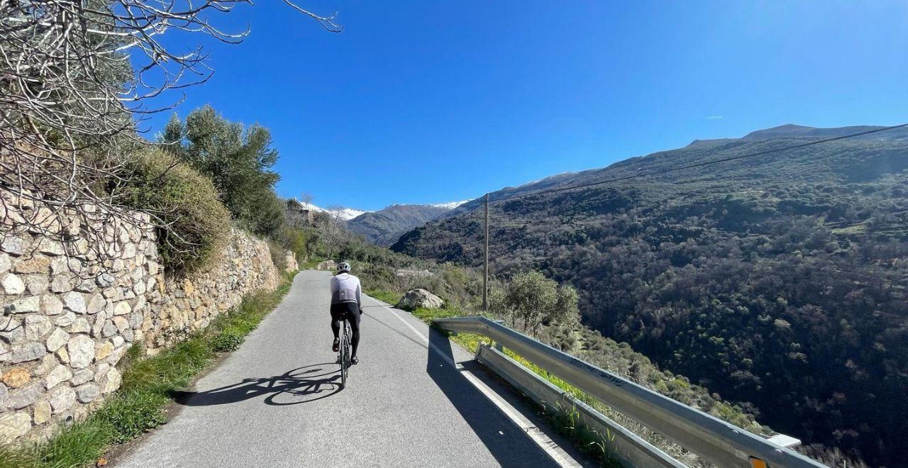 Cyclist riding on a narrow road through scenic mountains and stone walls under a clear blue sky.