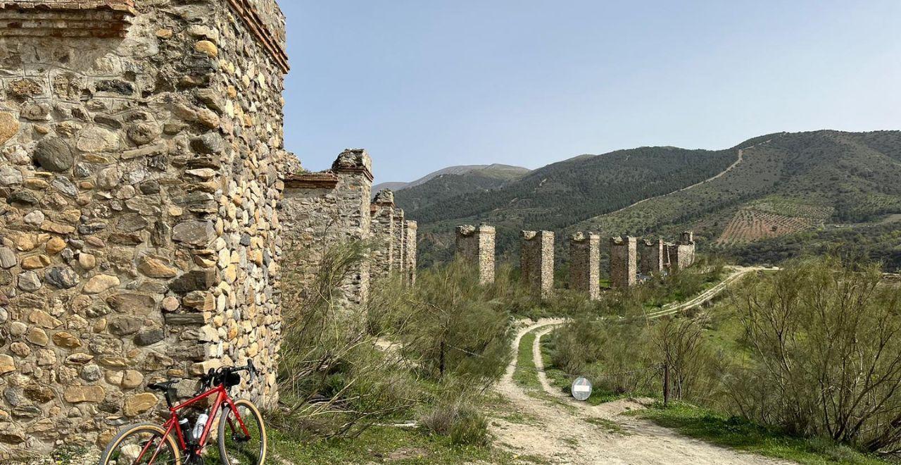 Red bike leaning against ancient stone aqueduct ruins with hills in the background.