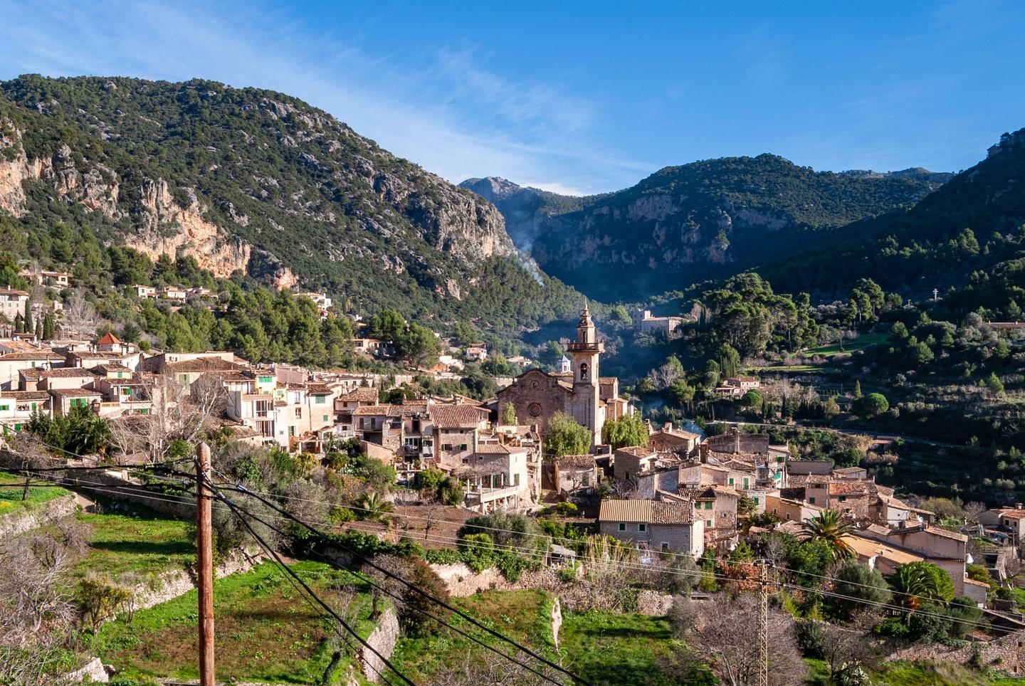Panoramic view of a quaint village nestled in the lush green mountains of Mallorca, featuring traditional stone houses and a prominent church tower under a clear blue sky.