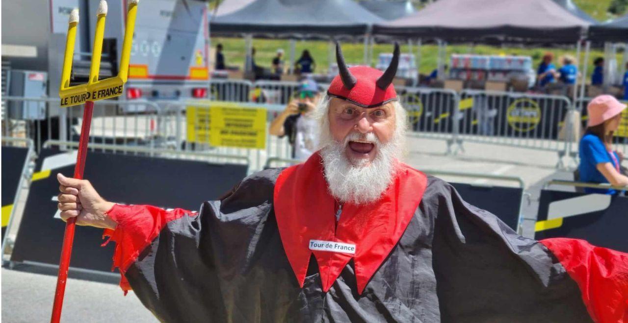 Cheerful elderly man dressed as the Tour de France devil, holding a trident and wearing a red and black costume at a cycling event