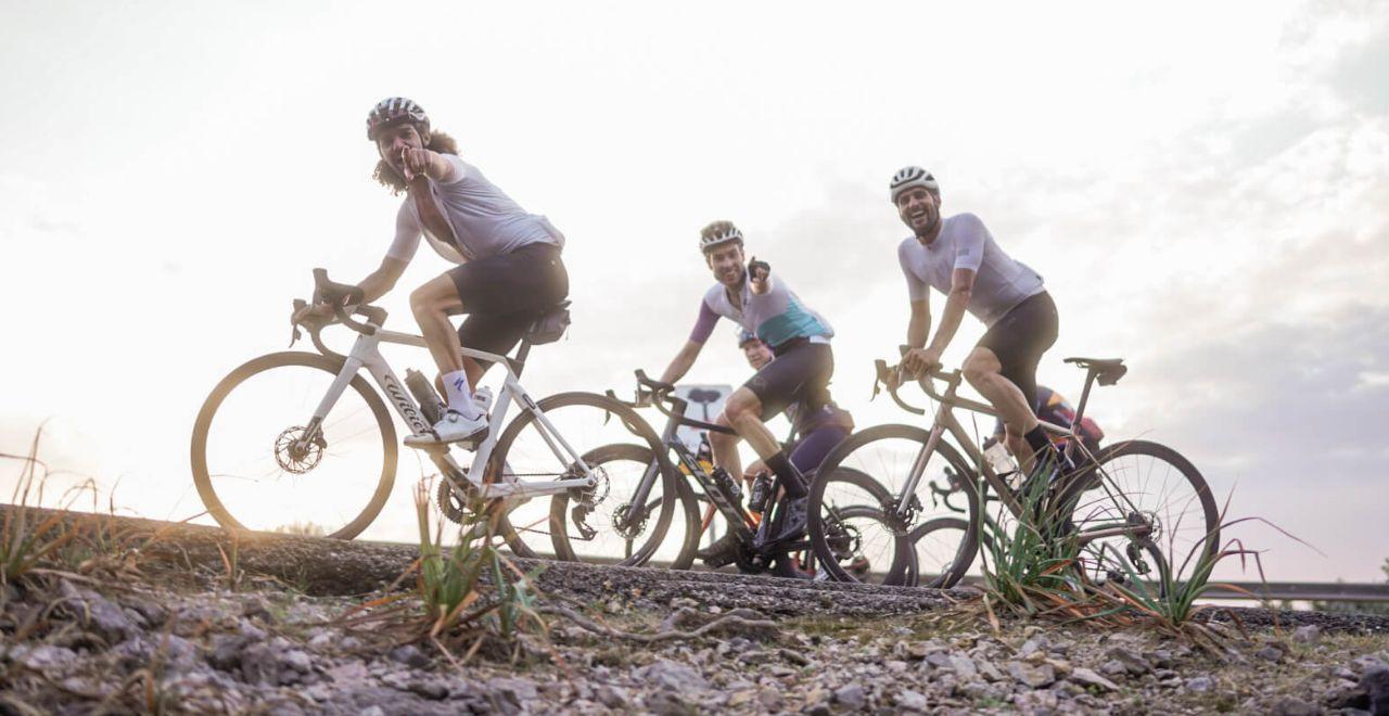 Group of enthusiastic cyclists pointing and smiling during a ride, with gravel terrain and an overcast sky in the background.