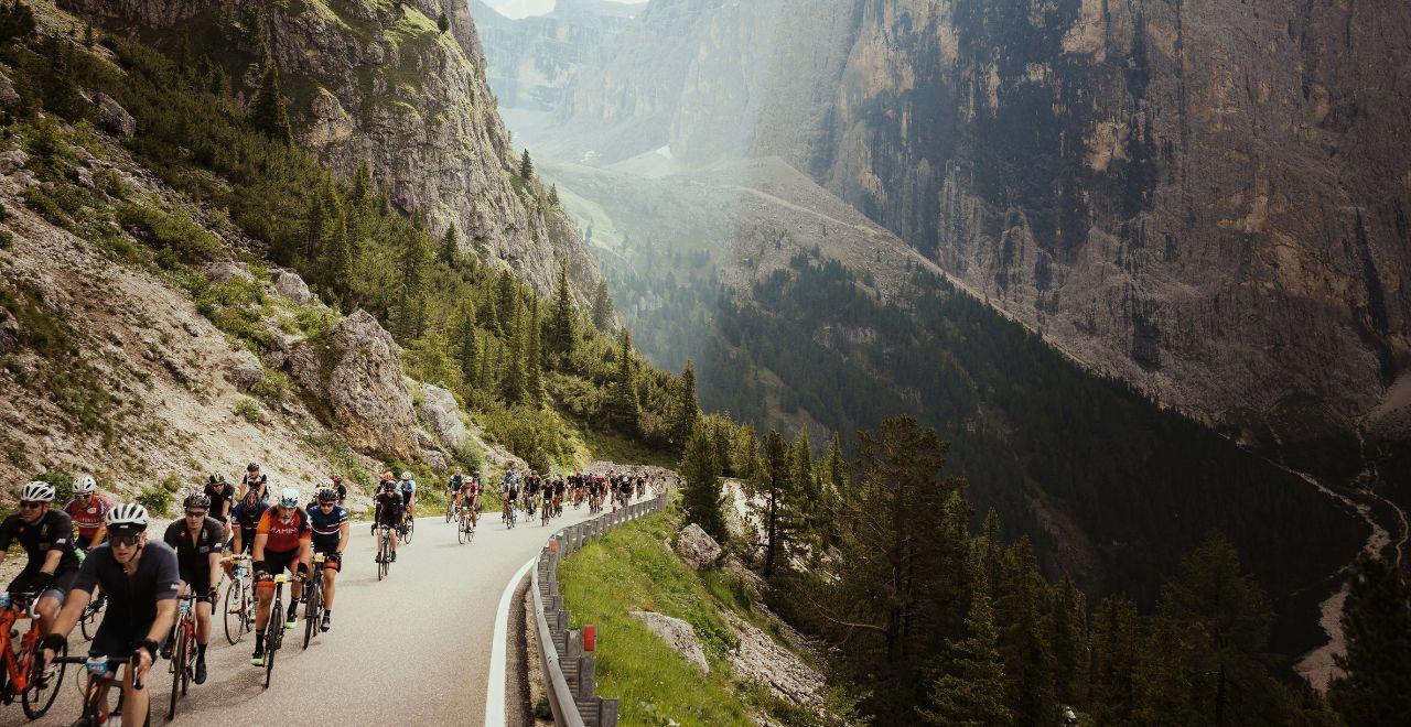 Cyclists riding along a narrow mountain road with towering cliffs and dense forest in the Maratona dles Dolomites