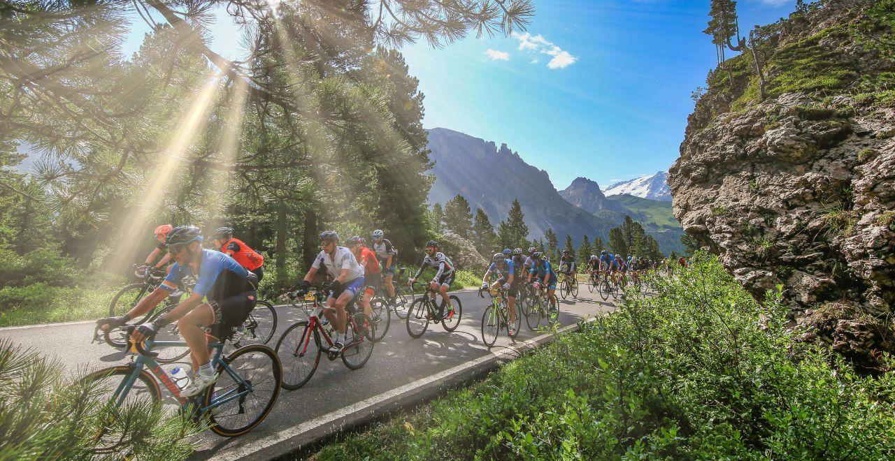 Cyclists riding through a sunlit forest with rays of sunlight breaking through trees during the Maratona dles Dolomites