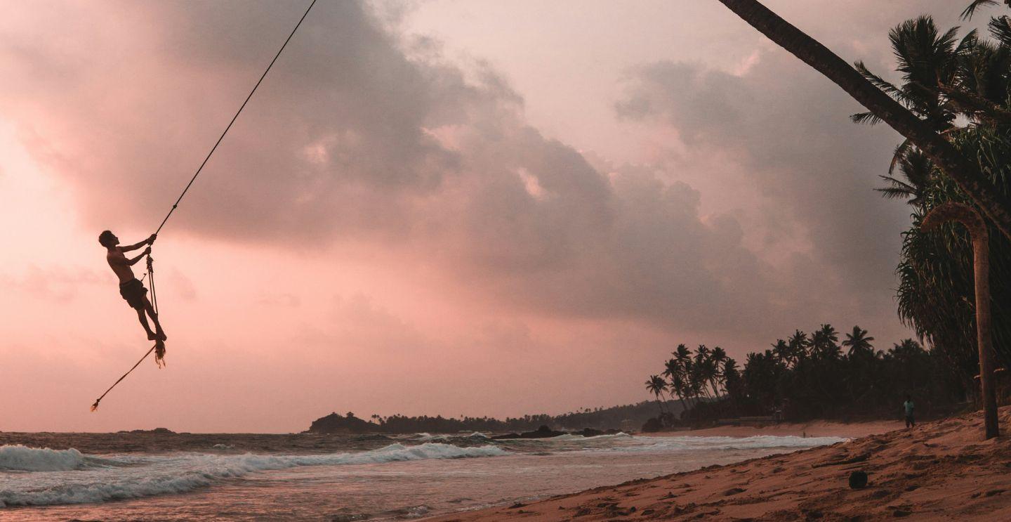 Person swinging on a rope tied to a palm tree on a beach at sunset with a pink sky and waves in the background.