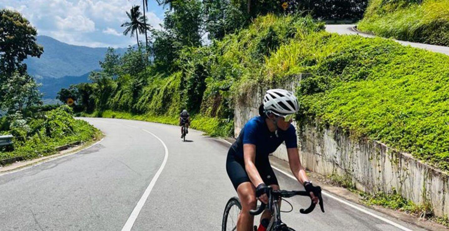 Cyclist in a blue jersey riding on a curving road through a green, mountainous landscape.