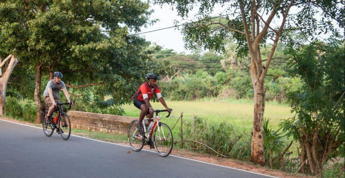 Two cyclists riding on a road surrounded by green fields and trees, with one cyclist wearing a red jersey.
