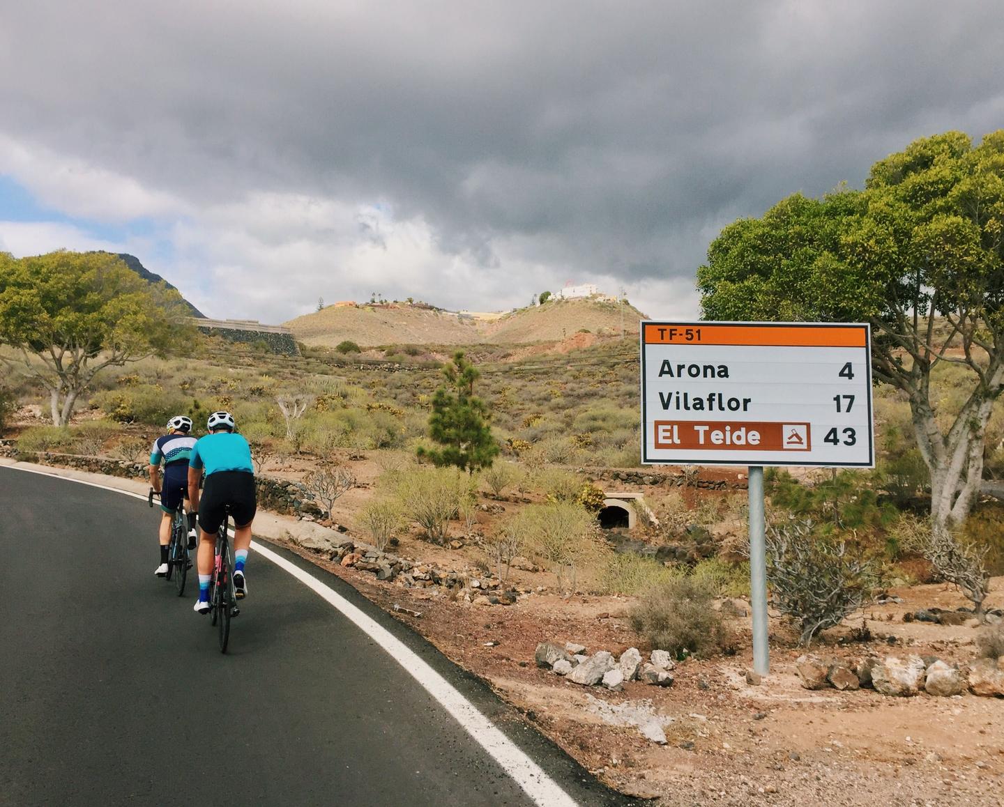 Two cyclists riding on a scenic road in Tenerife with a sign indicating distances to Arona, Vilaflor, and El Teide. The landscape features a mix of trees and hills under a cloudy sky.
