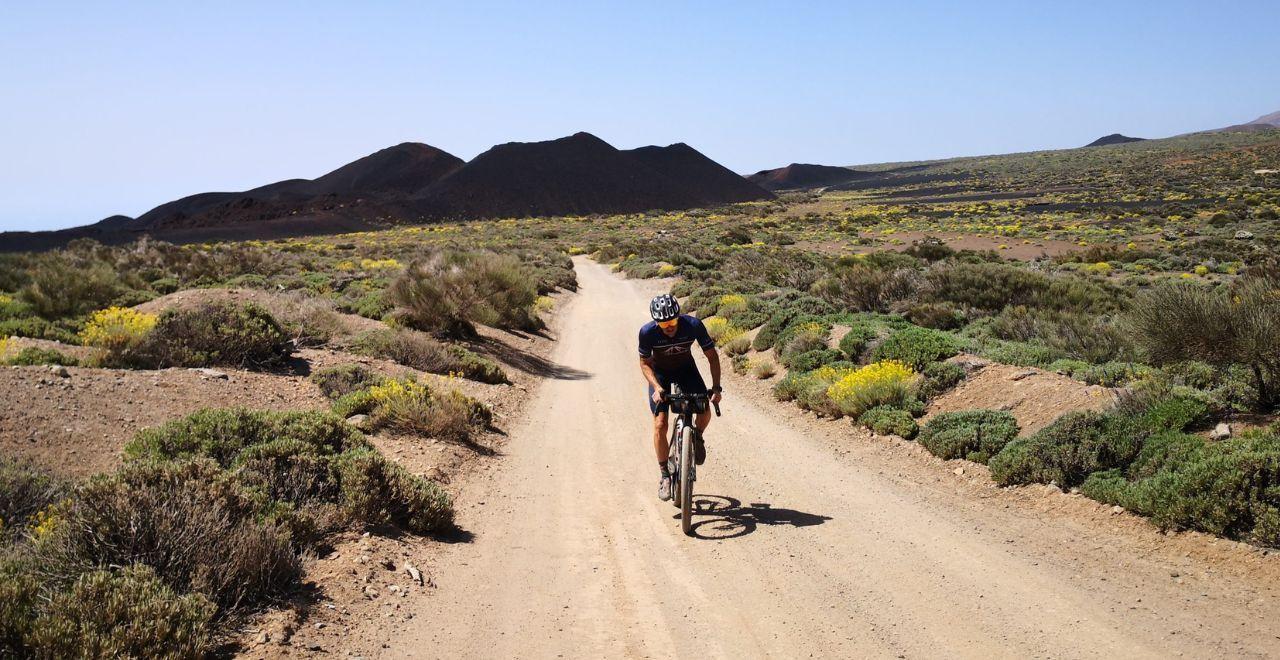 Cyclist riding on a sandy gravel path in Tenerife, surrounded by green shrubs and yellow wildflowers, with volcanic hills in the background