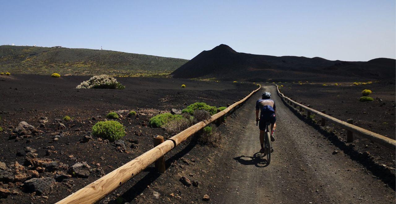 Cyclist riding on a gravel path through volcanic terrain in Tenerife, with sparse green vegetation and a distant hill under a clear blue sky.